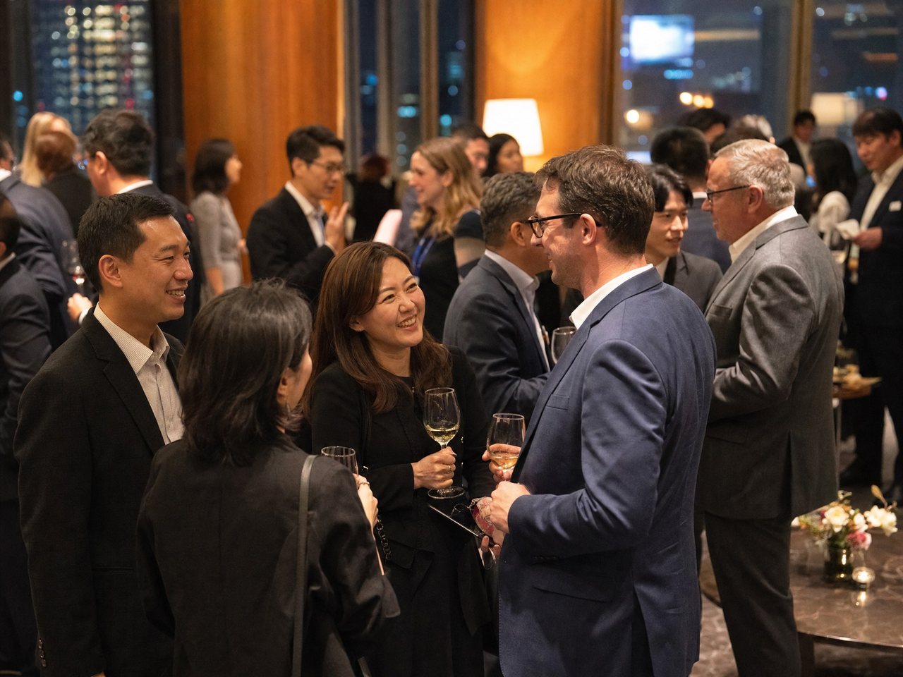 International researchers talking over drinks at an evening reception, Seoul skyline through the window